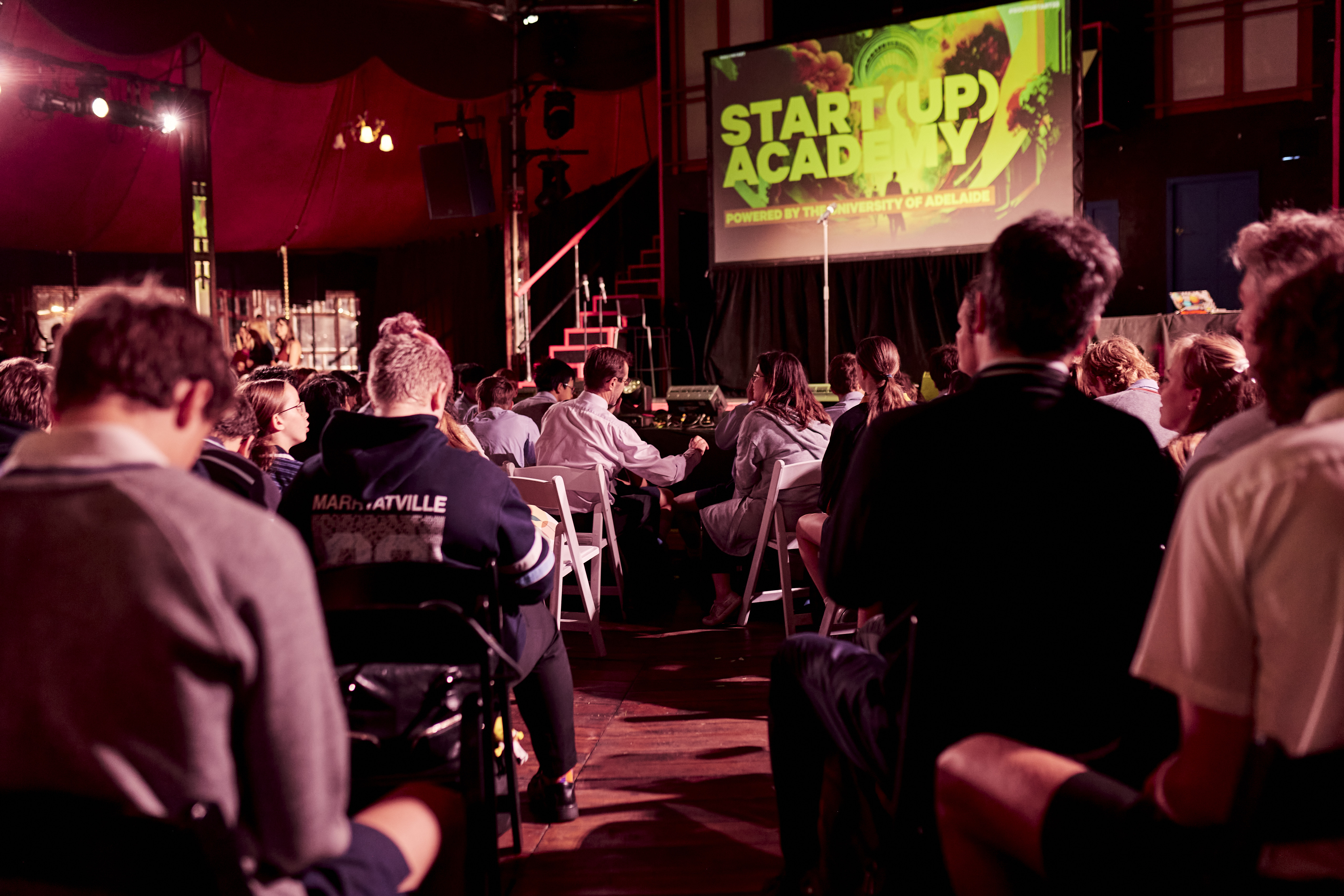 Students sitting inside an event tent, looking at a large presentation screen, waiting for the Startup Academy to begin.