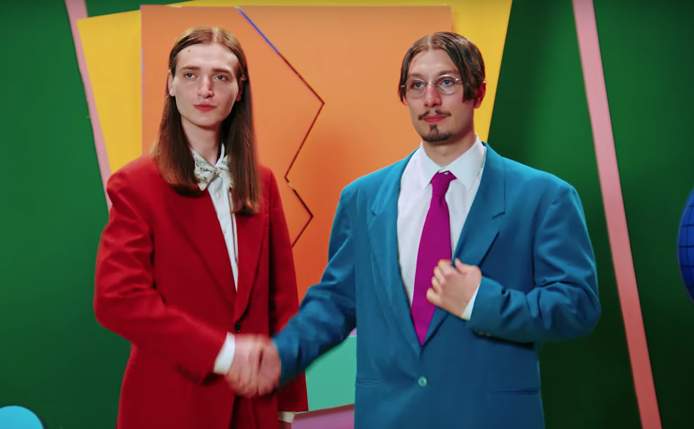 bbno$ and Y2K shake hands in a blue and red suit respectively.