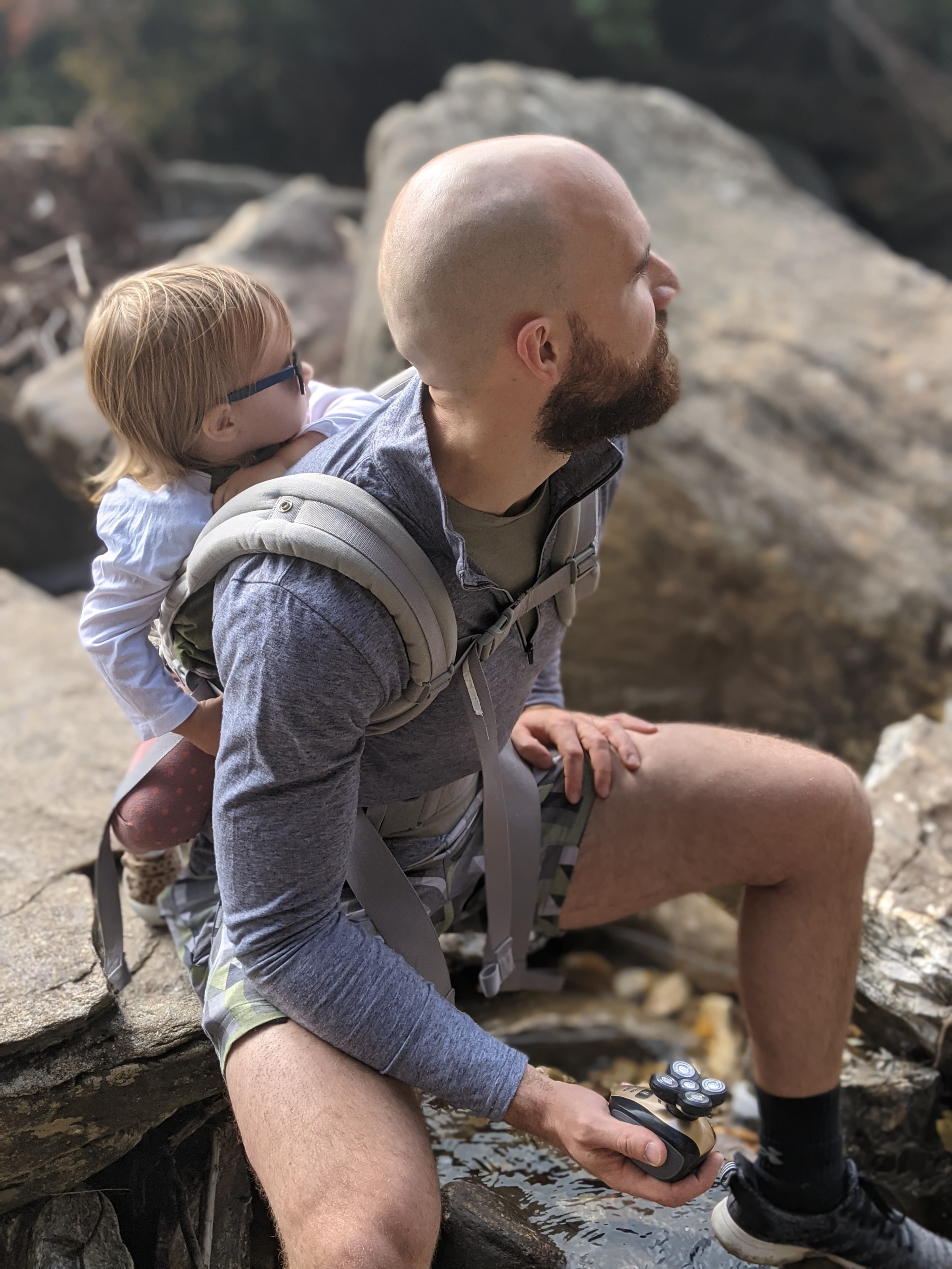  A bald male traveler carrying his toddler on his back enjoying the outdoors while holding a rotary electric shaver in his left hand 