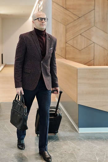 bald guy wearing glasses dressed in style while carrying a bag and pulling luggage at the airport