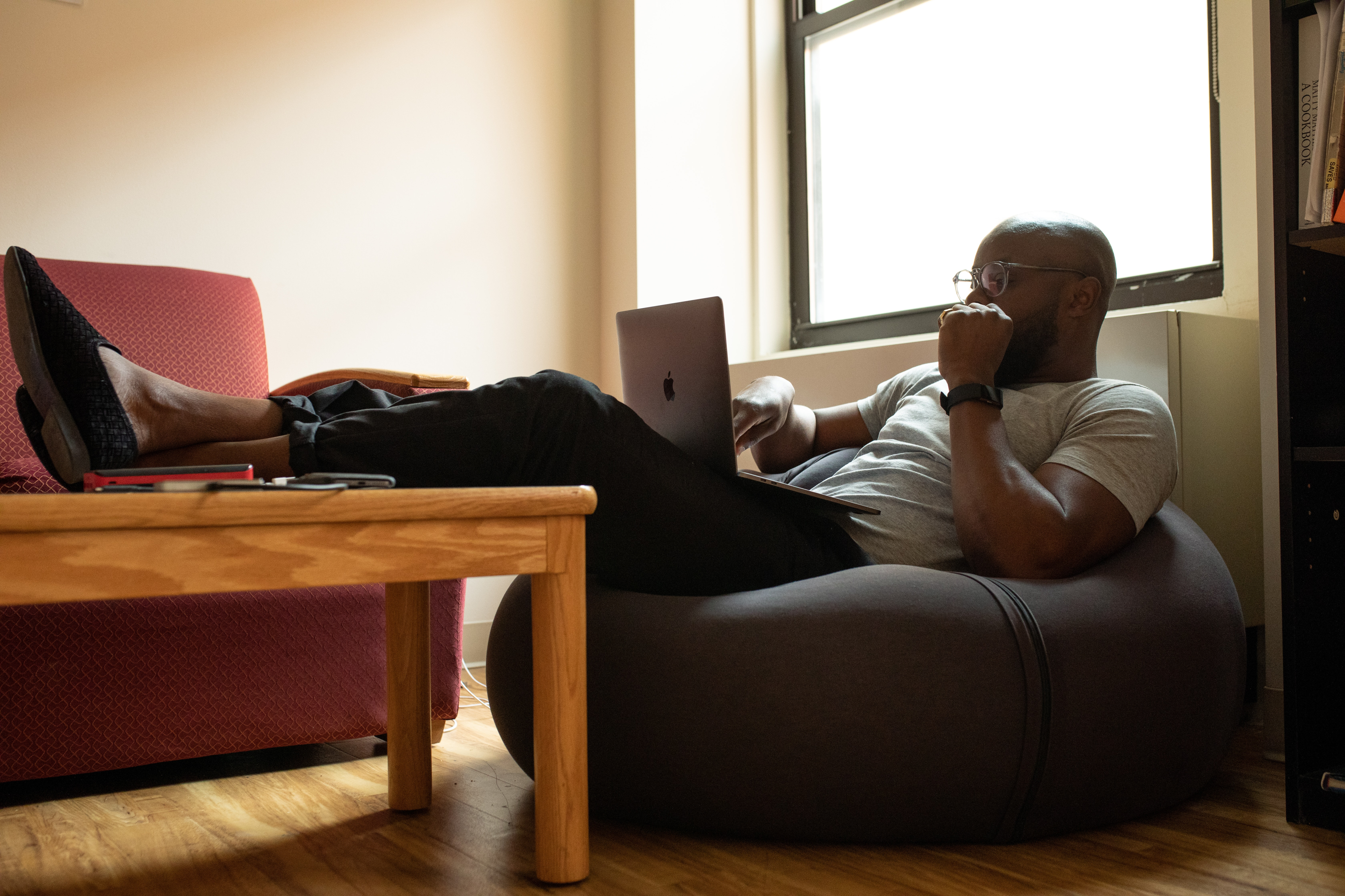 bald guy wearing glasses working on his laptop while sitting on a lazyboy and feet resting on a table